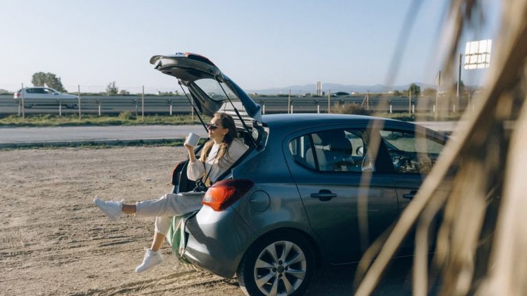 A woman sitting in the trunk of a parked car on a sunny day, enjoying a break during a road trip.